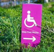 Image of pink wheelchair accessible sign in grass that says "Step free Route"